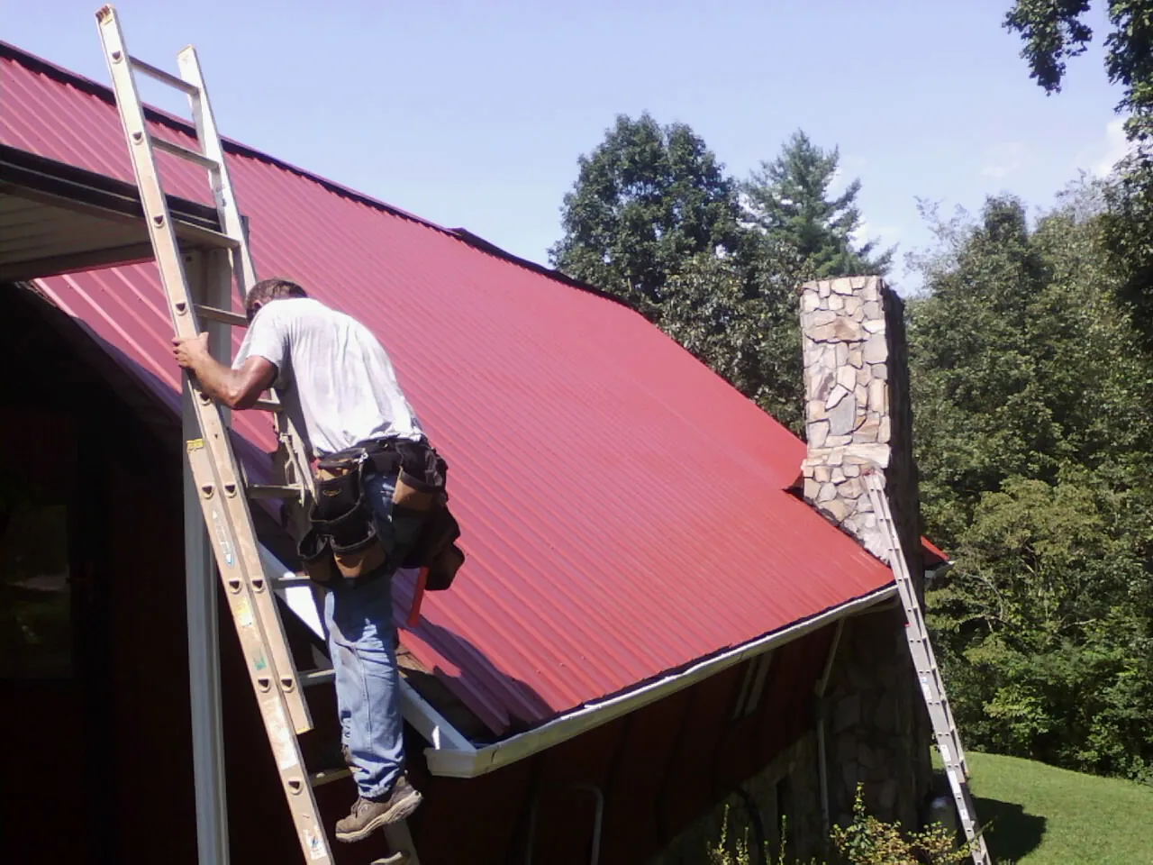 Roofer on a ladder beside a red metal roof and stone chimney in Cypress